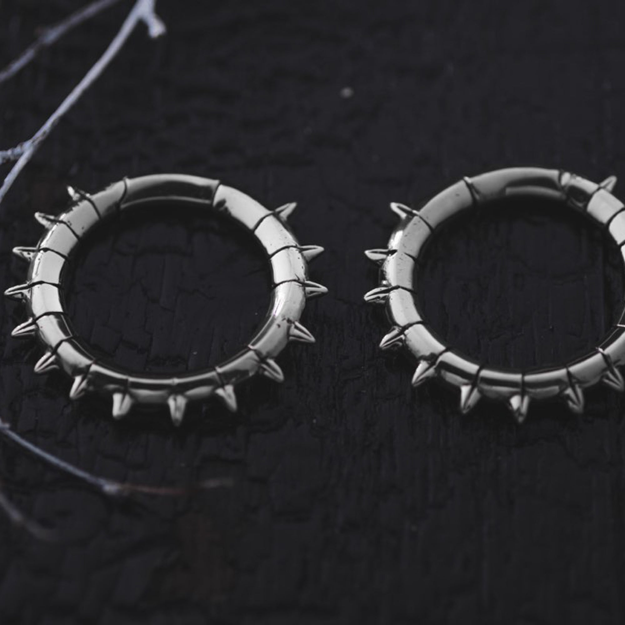 Close-up of a pair of Kaya Movement's KALASH Medium Spike Hoop Clicker Ear Weights in Silver Brass (4 gauge), adorned with small spikes around the edges, set against a textured black background. These tribal punk industrial jewelry pieces add a bold, edgy aesthetic to any outfit.